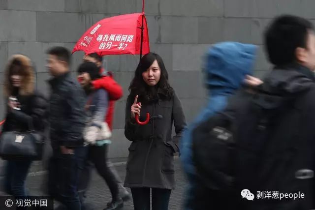 ▲2012年情人节，一家婚恋网站的模特撑开半把雨伞，上面写有“寻找风雨同路的另一半”的字样，站立在人流不息的上海南京西路街头。图片来自视觉中国