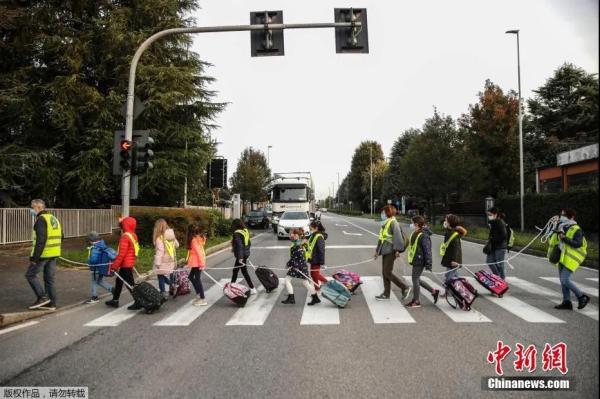 当地时间10月20日，在意大利北部贝卢斯科，孩子们拿着绳子保持社交距离，在志愿者的护送下走向学校。